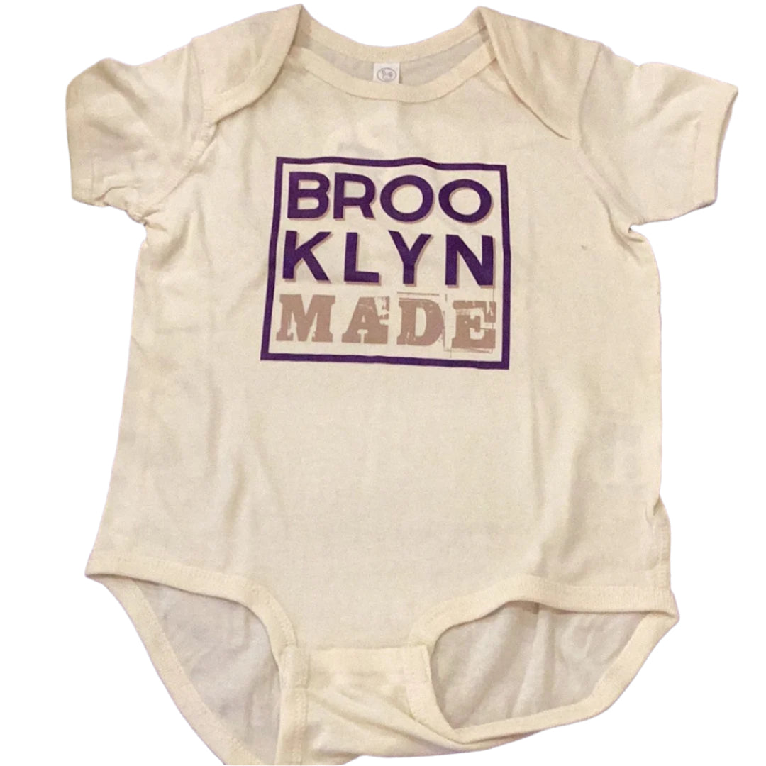 A white onesie with 'Brooklyn Made' printed in purple text on the front.