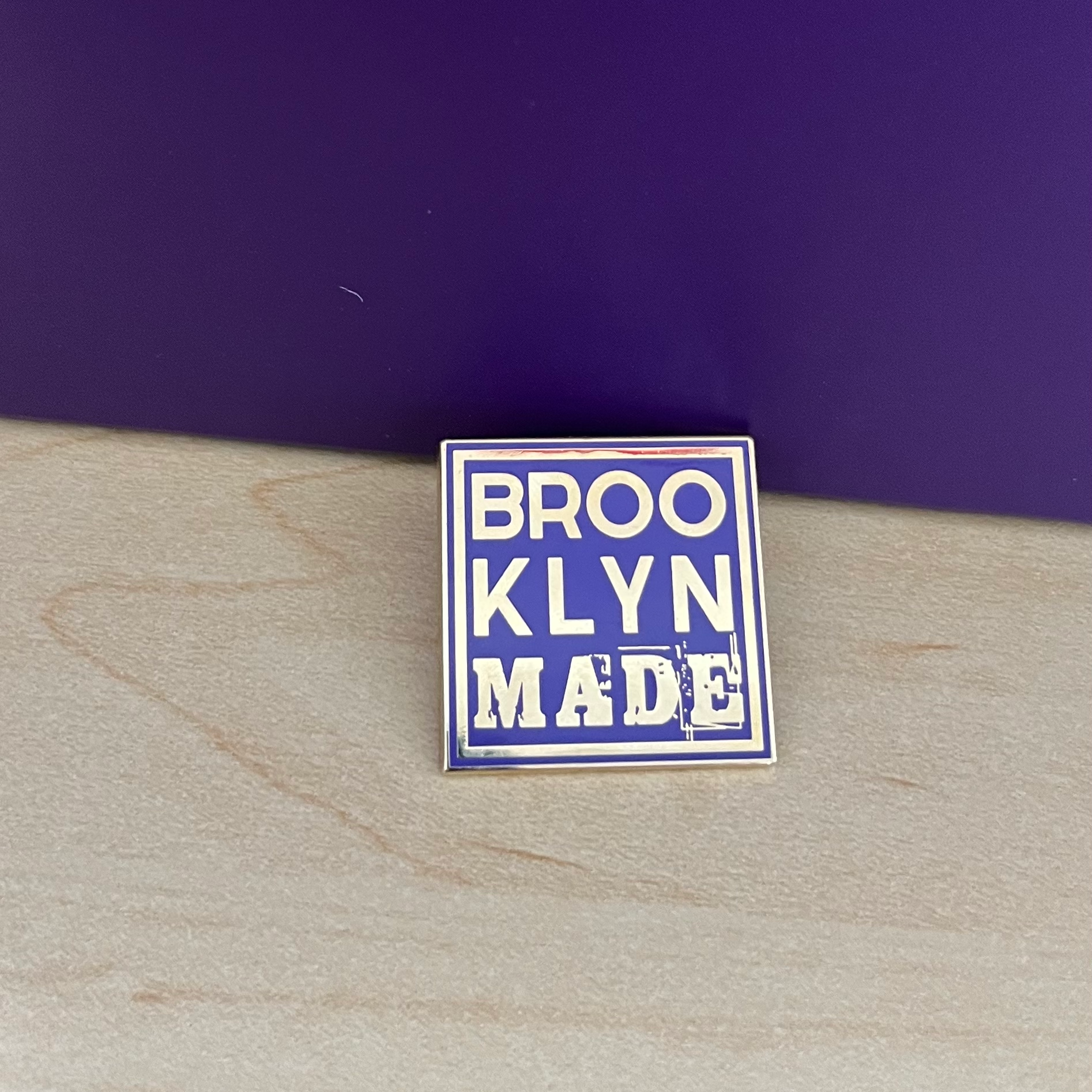 Enamel pin with 'BROOKLYN MADE' text on a wooden surface with a dark purple background