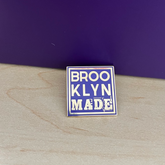 Enamel pin with 'BROOKLYN MADE' text on a wooden surface with a dark purple background