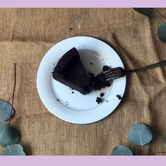 Chocolate cake on a white plate with a fork, on a textured brown surface with eucalyptus leaves.