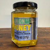 Jar of honey with a label reading 'Union St Honey Made by Brooklyn Bees' on a wooden surface.