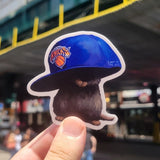 Hand holding a sticker of a person wearing a blue cap with a logo against a blurred street background