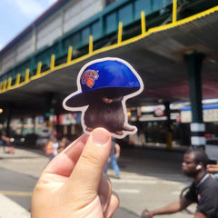 Hand holding a sticker of a person wearing a blue cap with a logo against a blurred street background